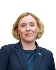 A person with shoulder-length blonde hair wearing a dark blue blazer and shirt against a plain white background.