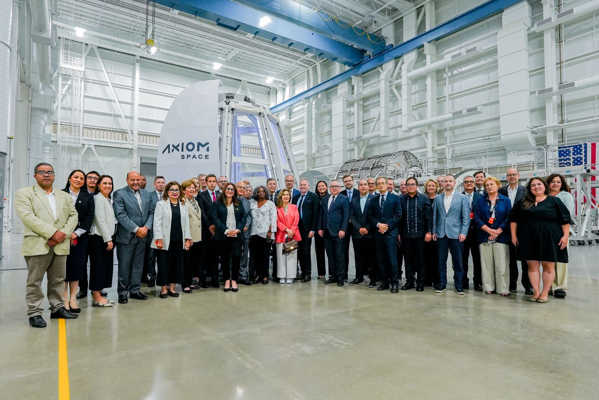 A group of professionals stands in a large industrial facility near an Axiom Space module, smiling for a group photo.