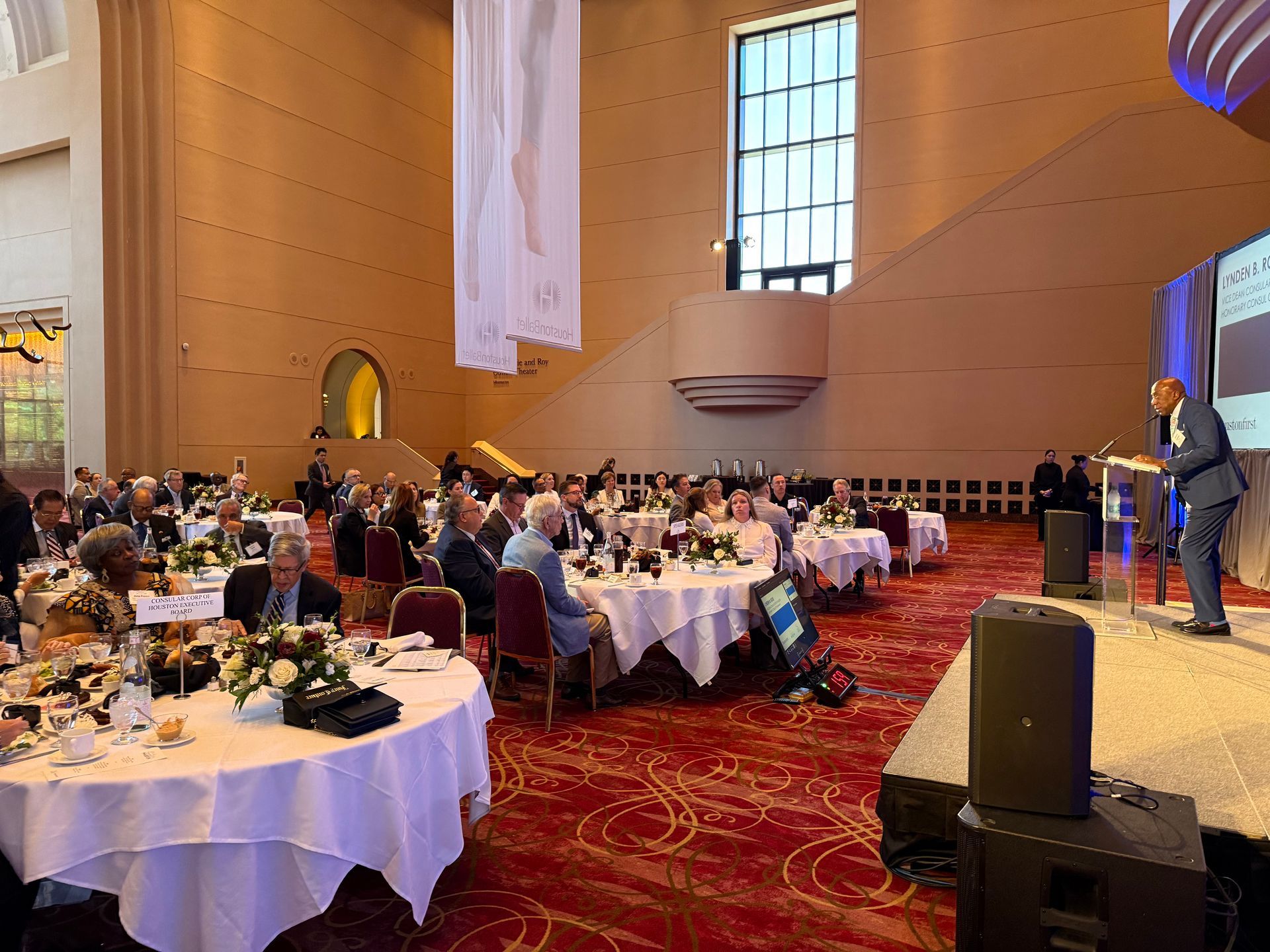 A speaker addresses a seated audience in a large ballroom with round tables, a red patterned carpet, and tall windows.
