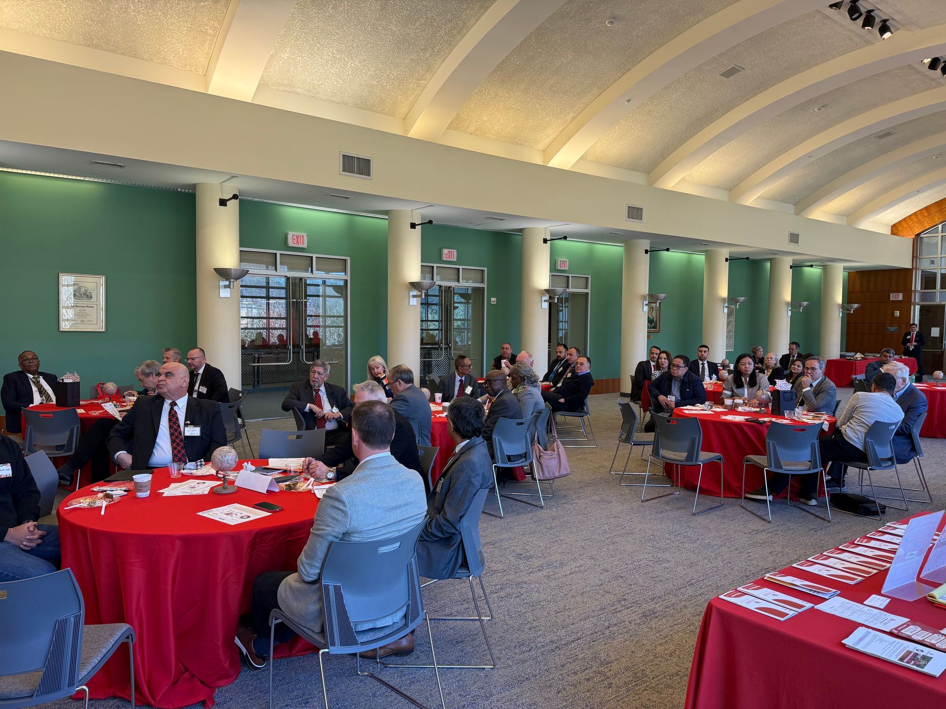 People in professional attire sit at round tables with red tablecloths during an indoor event in a large, airy hall.