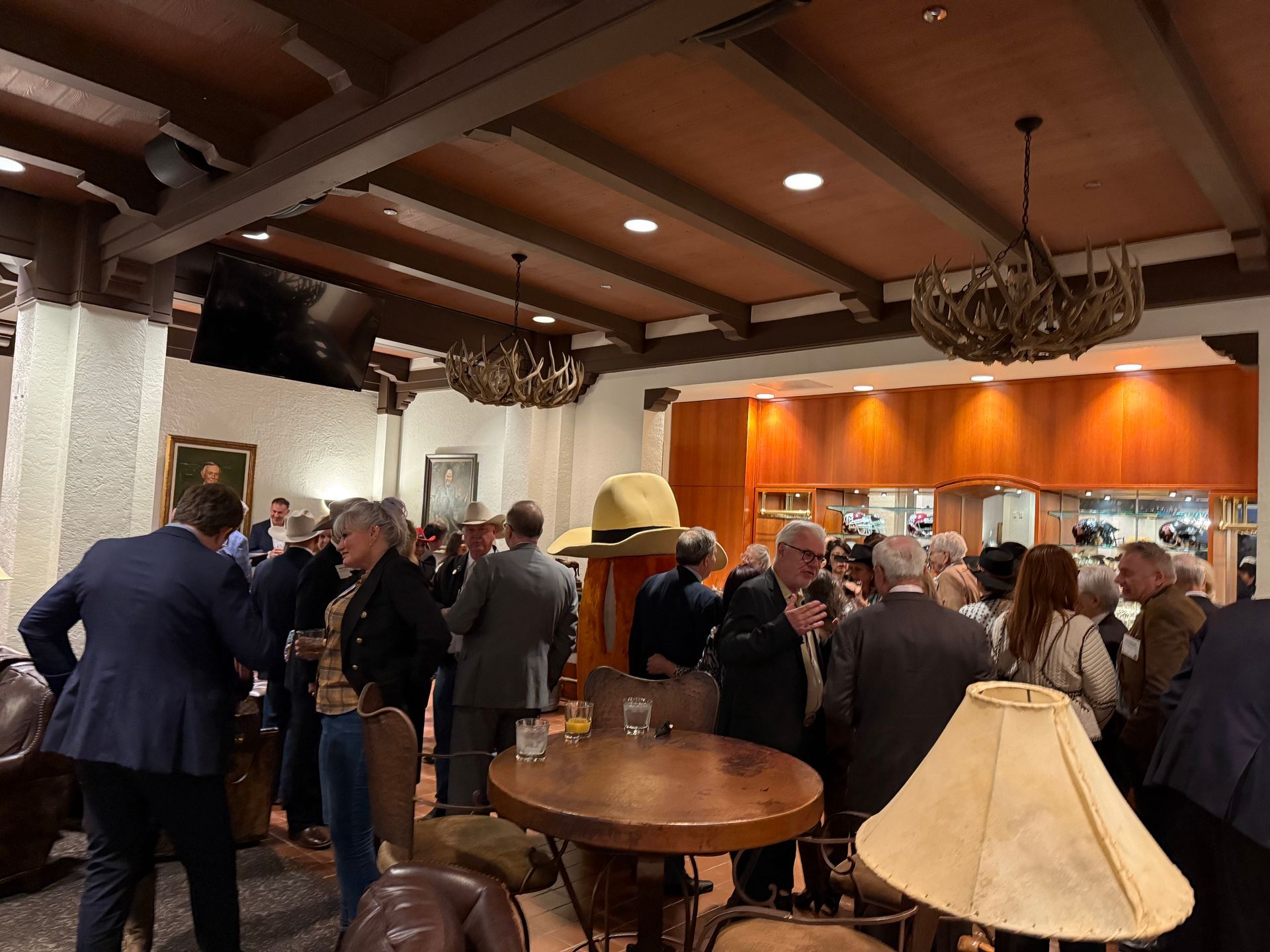 A group of people gathers for an indoor social event in a room with rustic wooden ceiling beams and antler chandeliers.