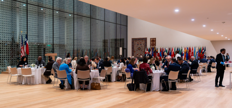 People dine at round tables in a large, brightly lit hall featuring a grid-patterned glass wall and rows of flags.