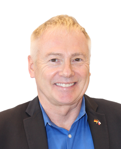 A smiling man wearing a blue shirt and a black blazer with a small pin on his lapel against a plain white background.
