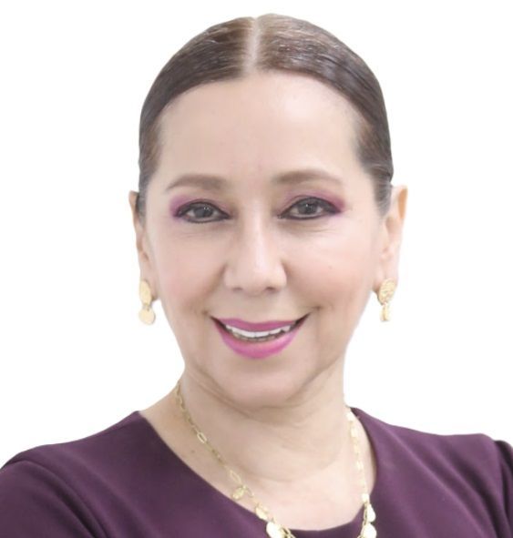 A smiling person with dark hair styled back, wearing a purple top, gold earrings, and a gold necklace against a white background.