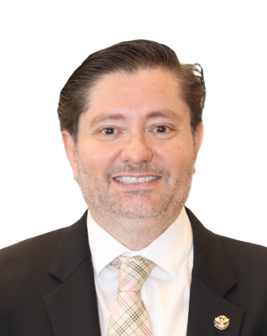 A person with brown hair and light facial hair wearing a dark suit, white shirt, and a plaid tie against a white background.