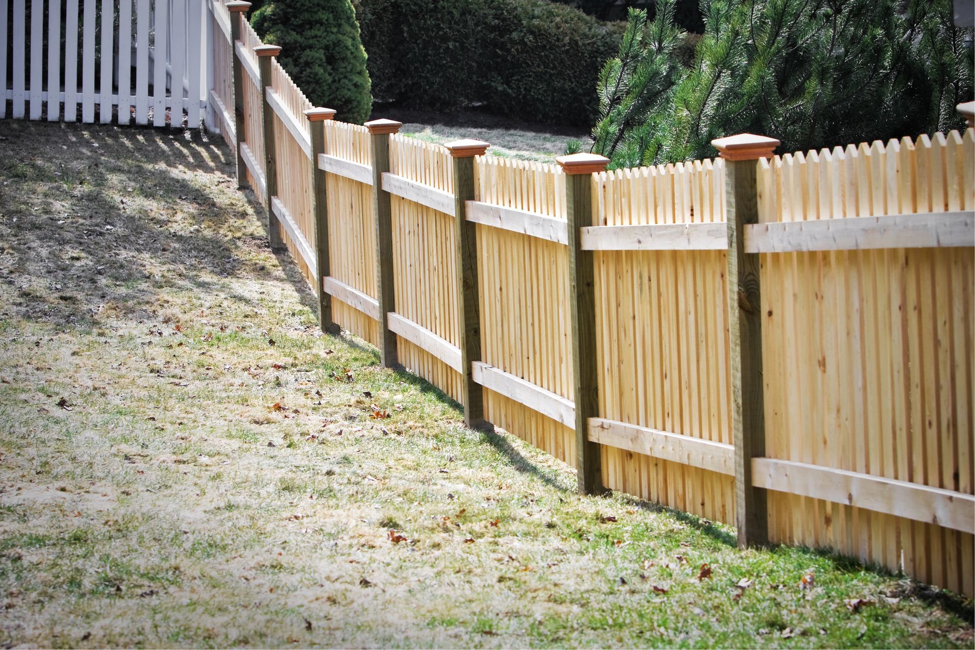 A wooden picket fence running along a grassy slope next to a white fence in a yard.
