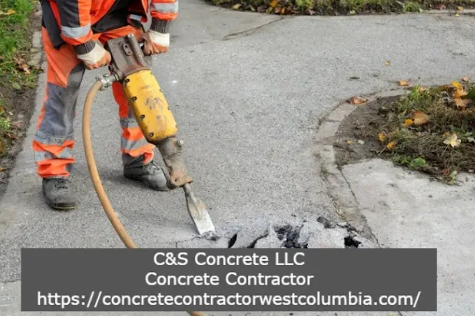 Worker in orange gear uses jackhammer on concrete, breaking it.
