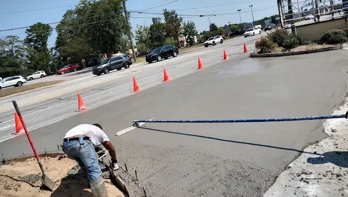 Construction worker smoothing wet concrete on a road, orange cones separate the work from traffic.