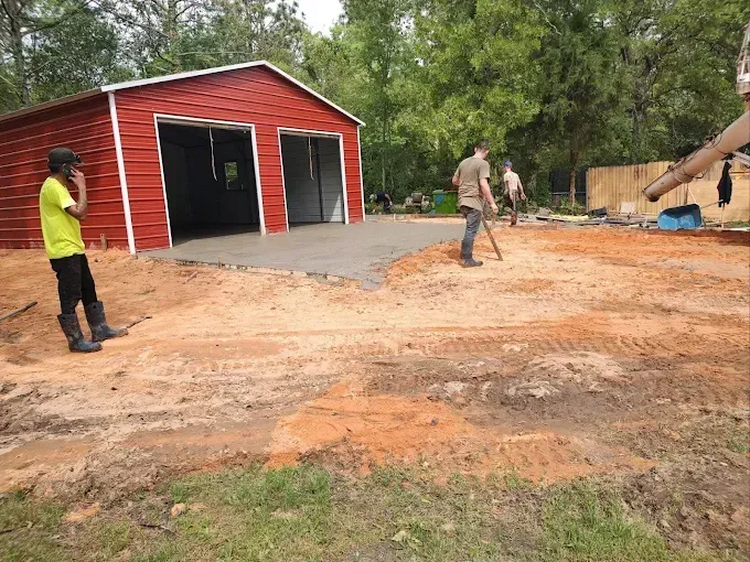 Workers pouring concrete for a garage driveway. Red garage, dirt ground, and trees in background.