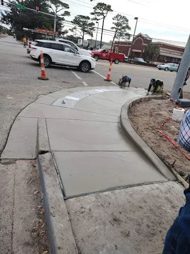 Construction of new concrete sidewalk at a street corner. Workers nearby. White SUV and red truck on street.