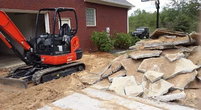 Orange excavator removing concrete slabs near a garage. Pile of broken concrete on the right.