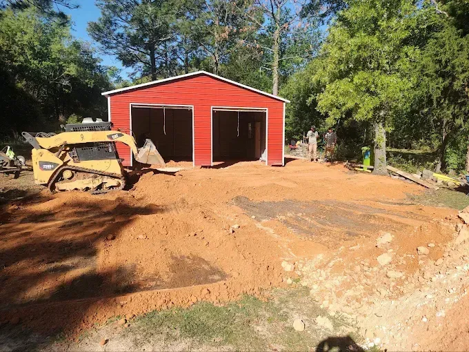 Red garage with two bays, construction site with earthmover, sunny day.