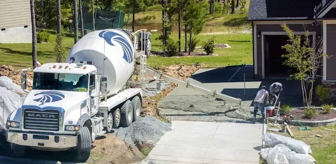 A cement truck pours concrete for a driveway under construction next to a house. A worker is smoothing the concrete.