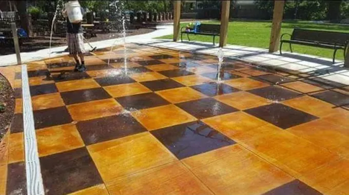 A person hosing a checkerboard-patterned concrete surface at a park, brown and tan squares, under a covered area.