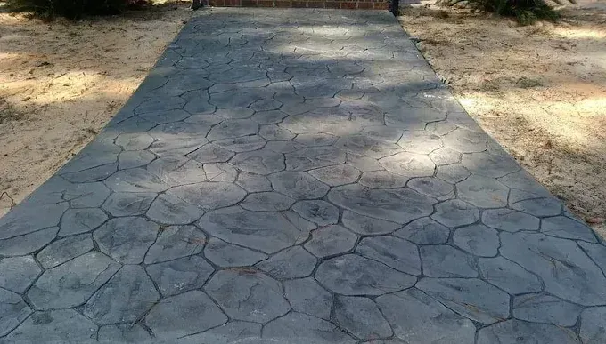 Gray concrete walkway with stone pattern, bordered by beige sand and trees.