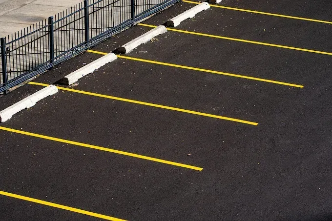 Empty parking lot with yellow lines and bumper stops along a dark paved surface next to a black fence.