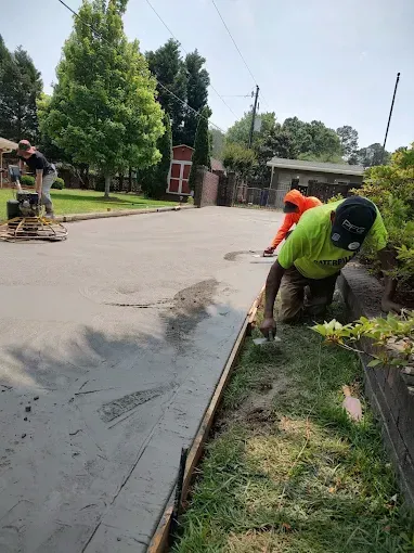 Construction workers laying concrete on a street, using tools; sunny day, green grass, trees.