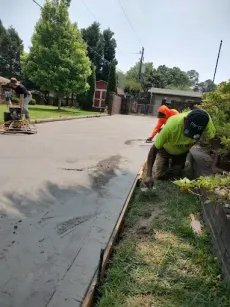Workers pouring concrete on a roadway. One kneels smoothing the edge, others stand nearby. Green, orange shirts.