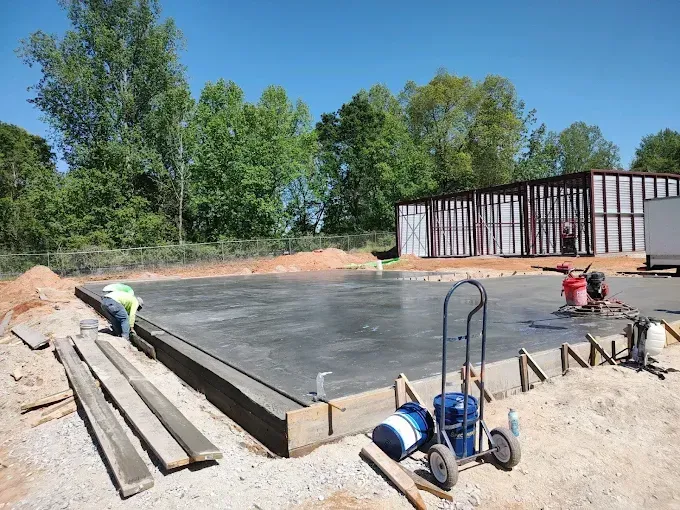 Concrete slab being poured at a construction site; workers, equipment, and a partially built structure are visible.