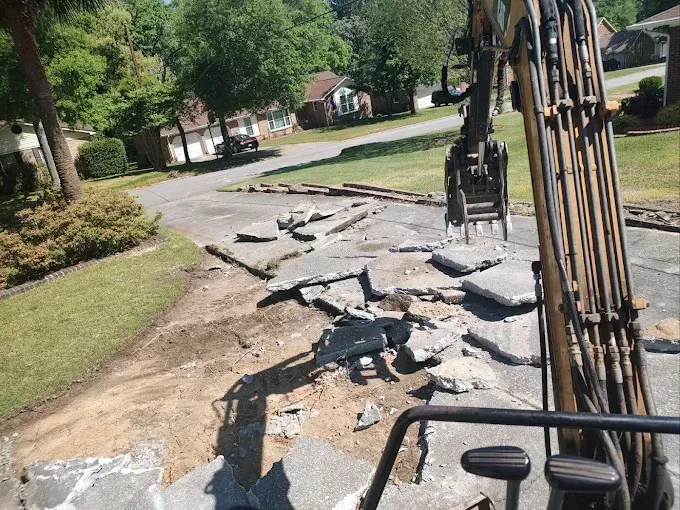 Excavator demolishing a driveway, breaking concrete into chunks on a sunny day.