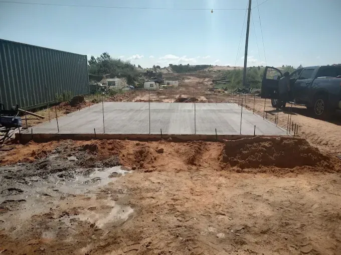Concrete slab under construction in a dirt lot. Rebar, soil, and a truck are visible.