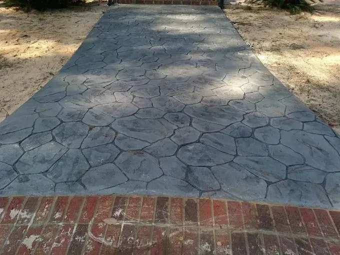 Gray stone-patterned concrete walkway bordered by red brick, surrounded by sand and trees.
