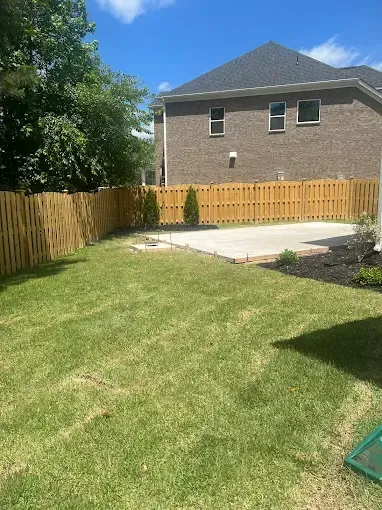 Backyard with a wooden fence, concrete patio, grass, and a brick house under a blue sky.