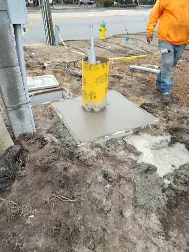 Construction site: concrete base with yellow cylinder, worker in orange jacket nearby.
