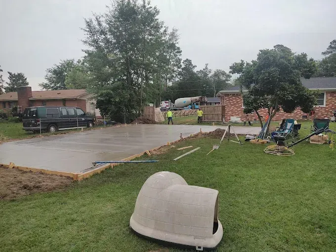 Concrete slab being poured in a residential backyard, dog house in foreground, workers and concrete truck in background.