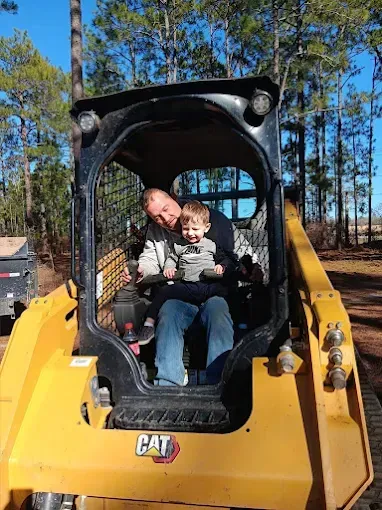 Man and young child sitting inside a yellow CAT skid steer; child smiles. Outdoors, sunny.
