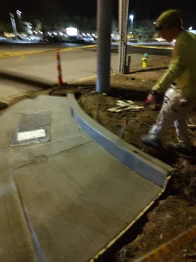 Construction worker near a concrete sidewalk and curb. Worker in light green shirt, gray curb, night setting.