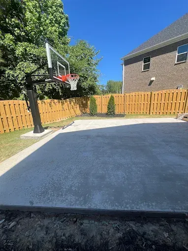 Basketball hoop on a concrete court in a backyard, with a wooden fence and a house in the background.
