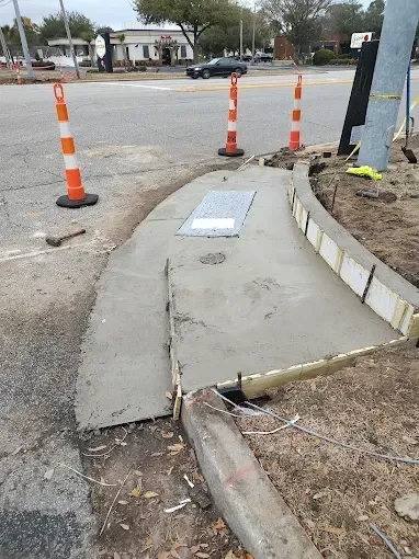 Fresh concrete sidewalk construction; orange cones, wooden forms, utility access panel, near a street.