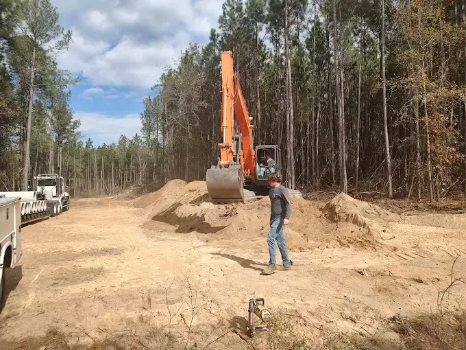 An excavator moving dirt in a clearing in a wooded area; a person stands nearby.