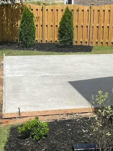 Newly poured concrete patio with wooden borders, set in a backyard with landscaping and a wooden fence.
