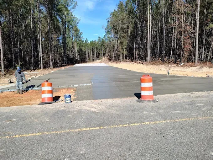Road construction: A worker near orange barrels, a newly poured concrete section, and a tree-lined background.