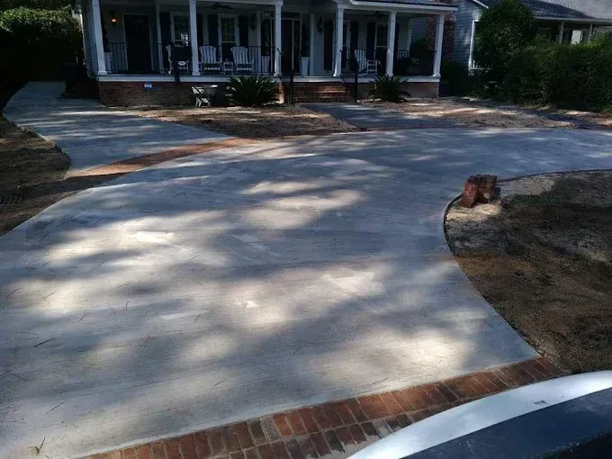 Concrete driveway curving toward a house with brick edging and surrounding landscaping.