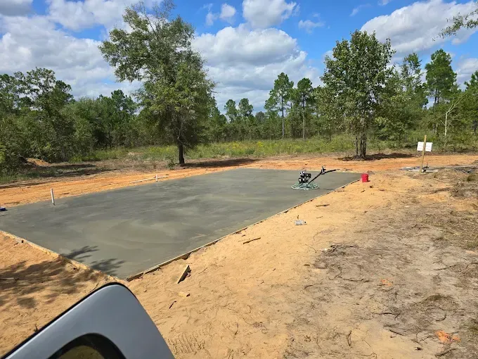 Newly poured concrete foundation in a sandy clearing, surrounded by trees under a blue sky.