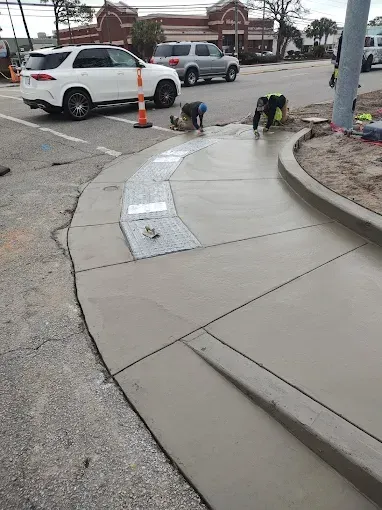 Construction workers pouring concrete for a sidewalk at a street corner. White car, gray concrete, and a brick building in the background.