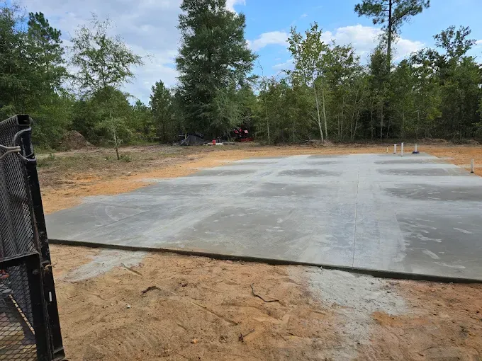 A freshly poured concrete pad on a dirt lot, surrounded by trees under a blue sky.