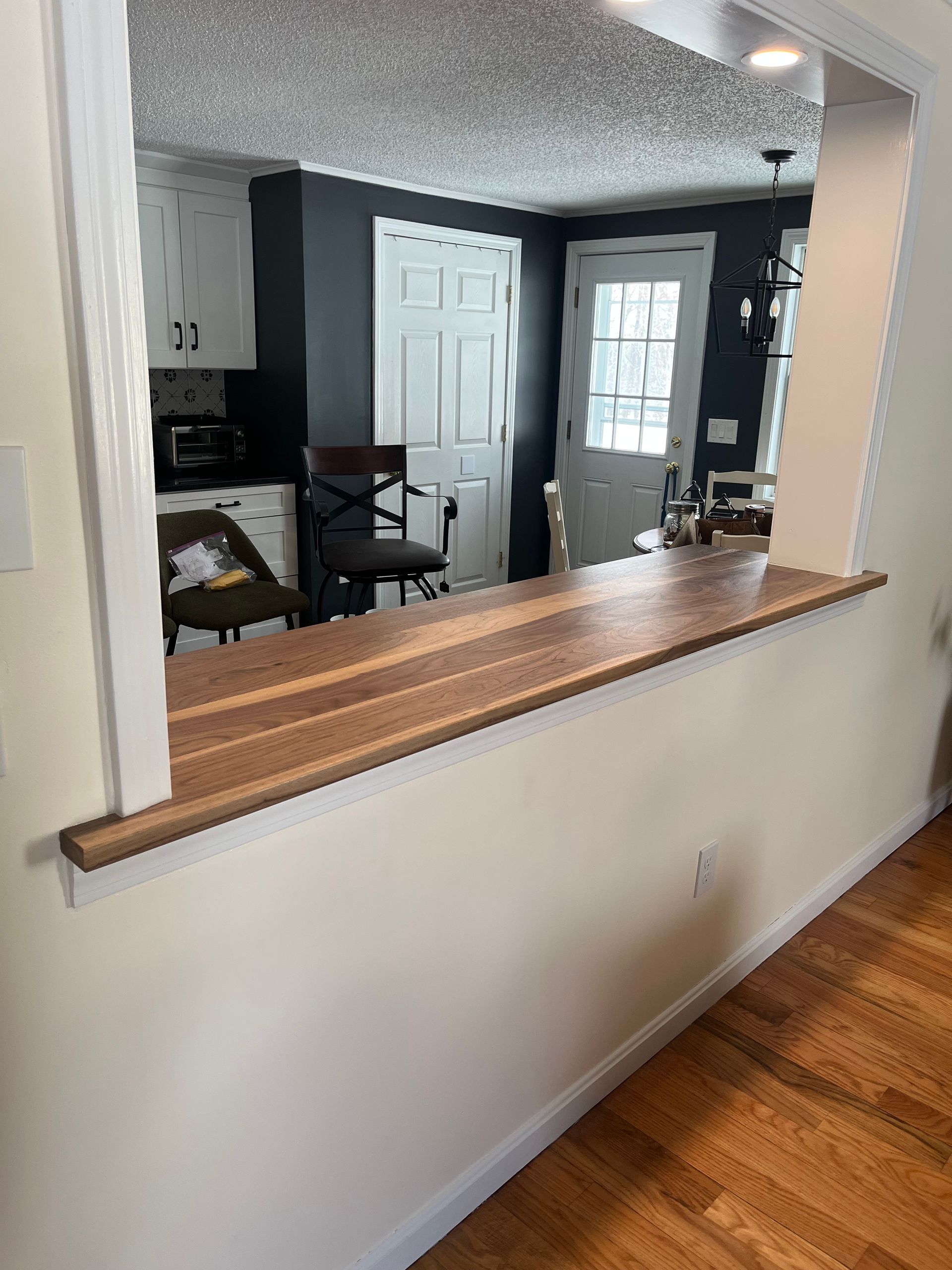 A wooden countertop spans a kitchen pass-through window looking into a kitchen with dark grey walls and white cabinets.