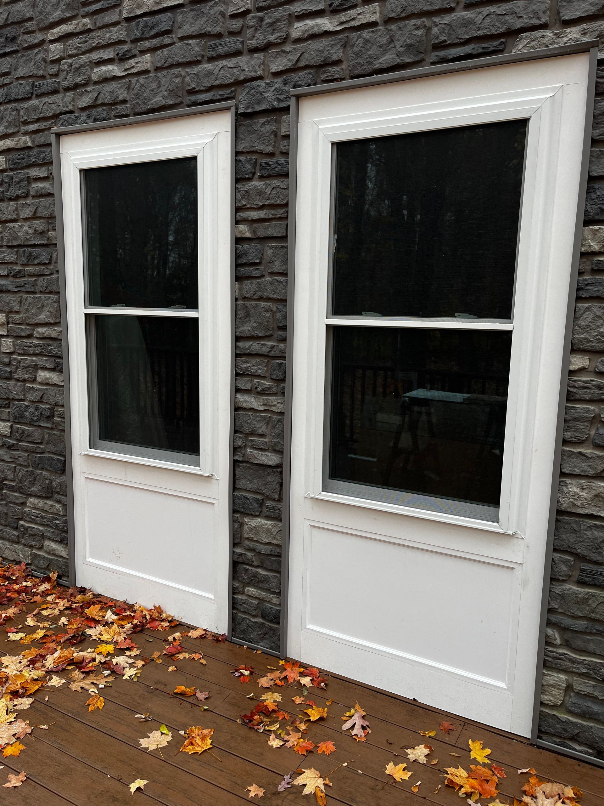 Two white storm doors with glass panes installed on a dark stone exterior wall above a wooden deck covered in fallen leaves.