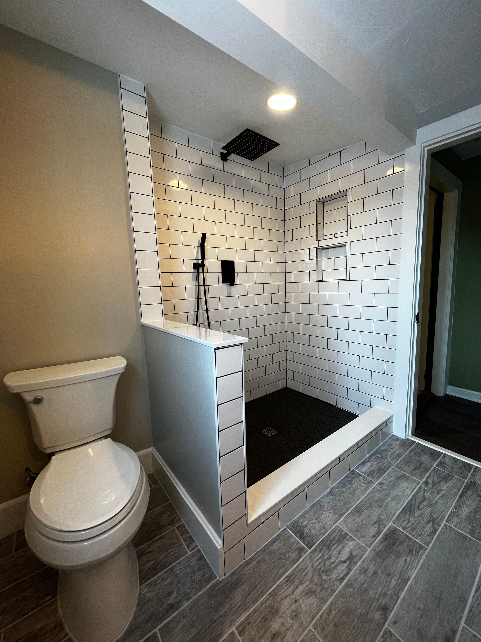 A bathroom featuring a walk-in shower with white pixelated tiles, a black floor, and a nearby white toilet on gray flooring.
