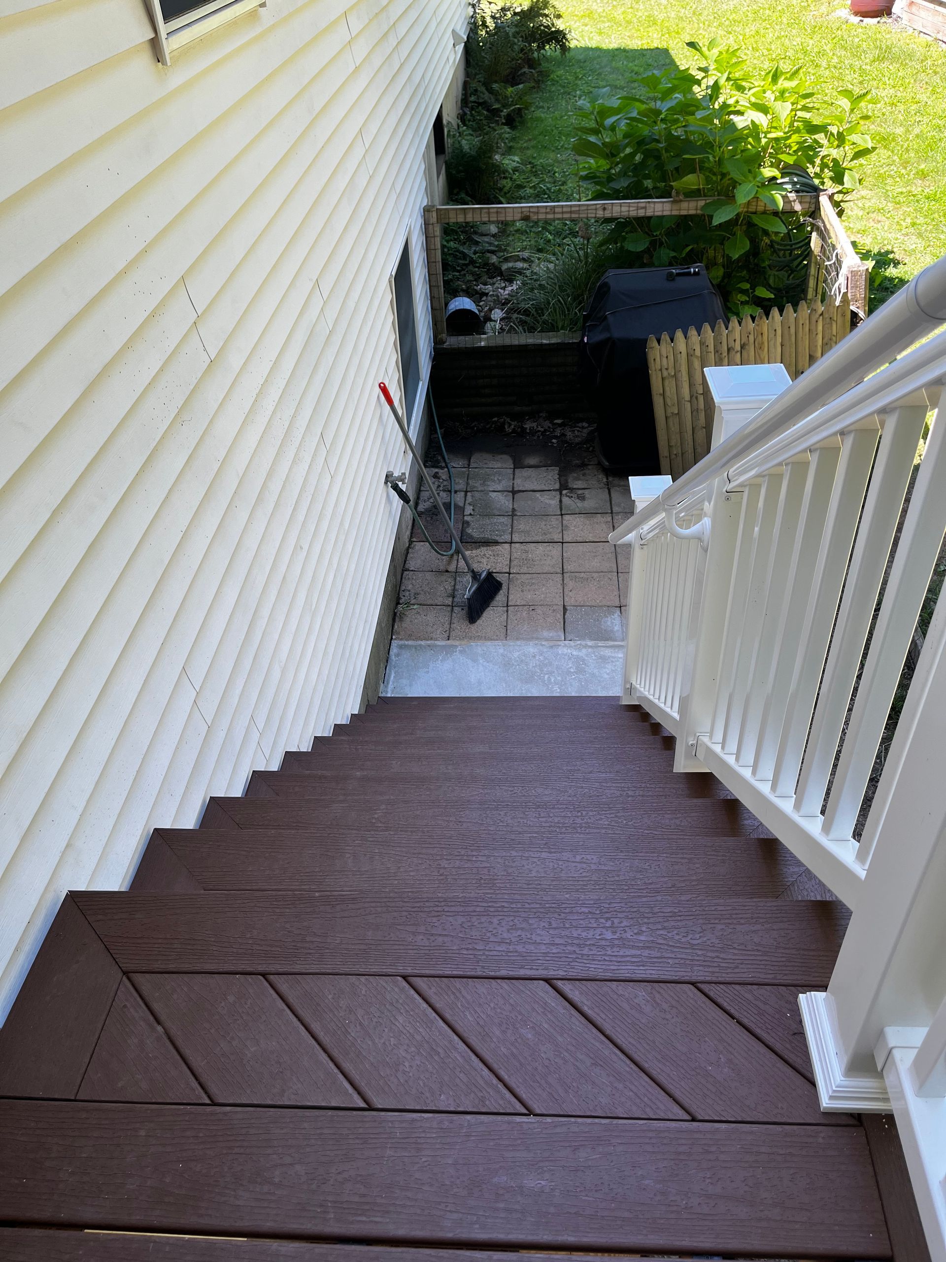 A high-angle view of brown deck stairs leading down to a paved backyard area next to a light-colored house.