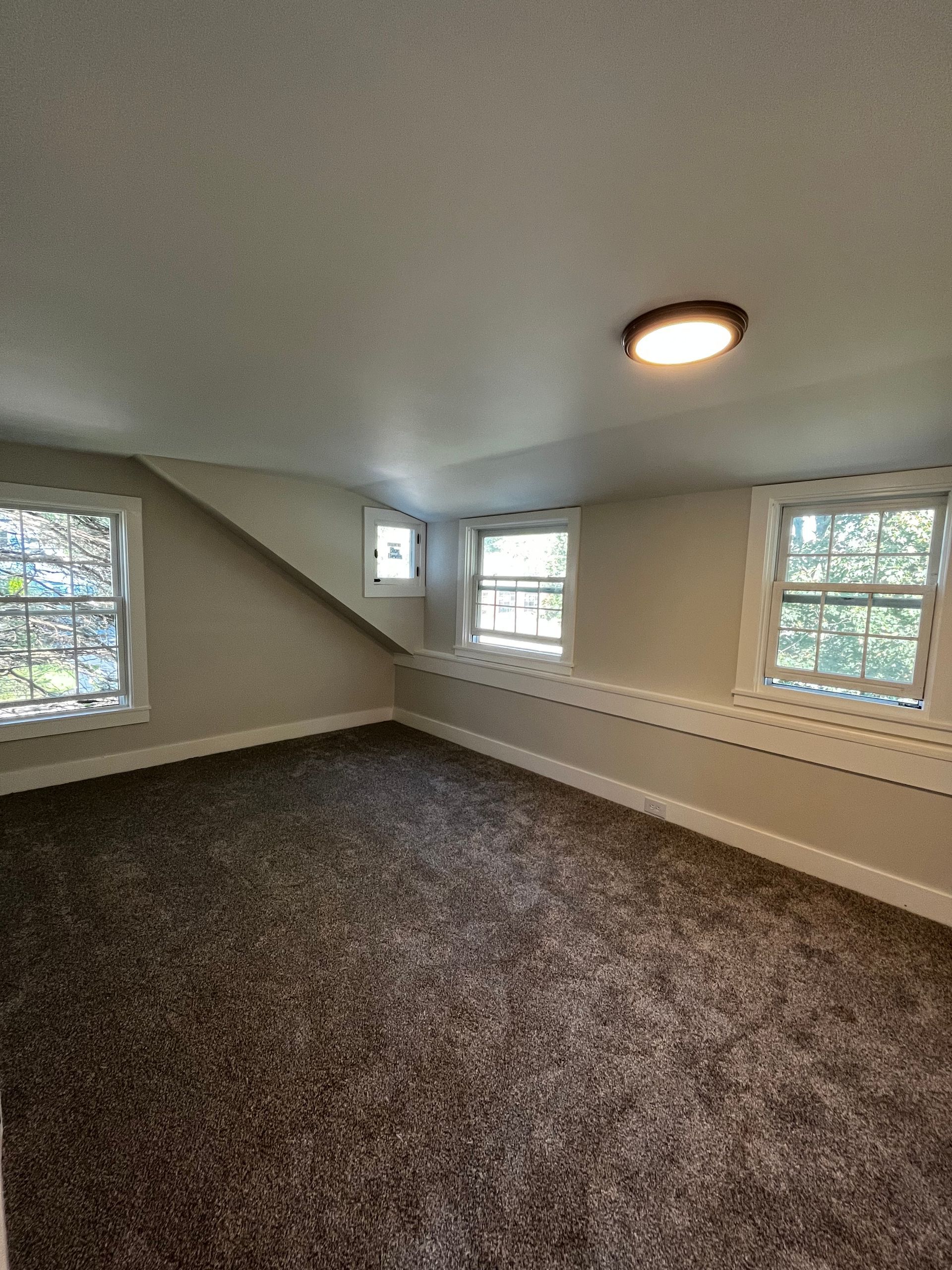 An empty carpeted attic room with slanted walls, two windows, a small side window, and a ceiling light.