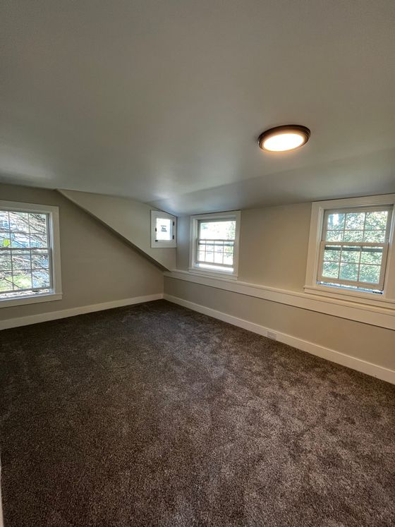 An empty carpeted attic room with slanted walls, two windows, a small side window, and a ceiling light.