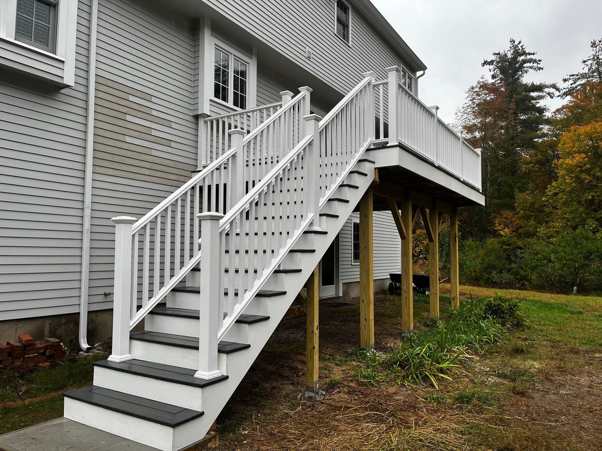 A raised deck with dark-colored stairs and flooring, white railings, and light-colored siding, situated near a forest.