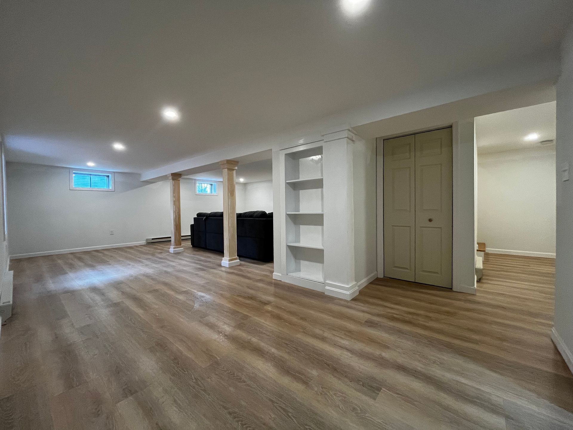 A bright, finished basement with light wood-look flooring, white walls, built-in shelving, and a black couch in the back.