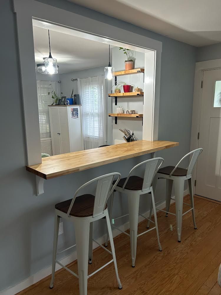 A pass-through kitchen counter with three metal bar stools set against a gray wall, featuring wooden shelving inside.