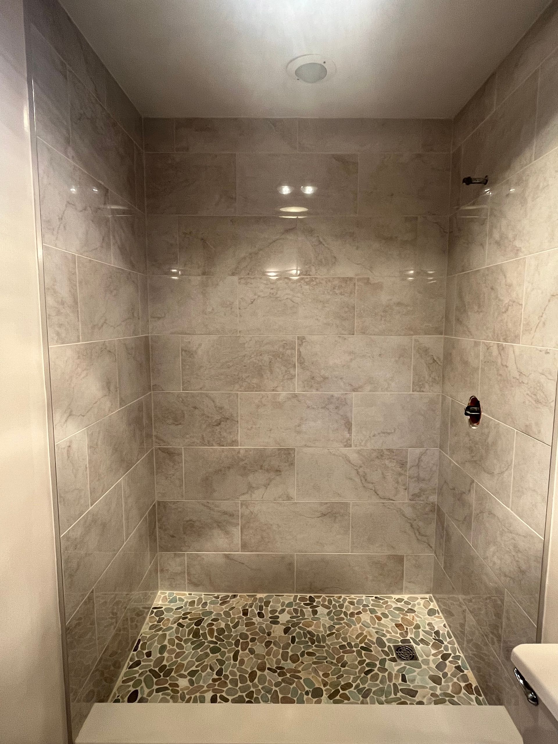 A walk-in shower with large, light-colored rectangular wall tiles and a textured, multi-colored pebble tile floor.